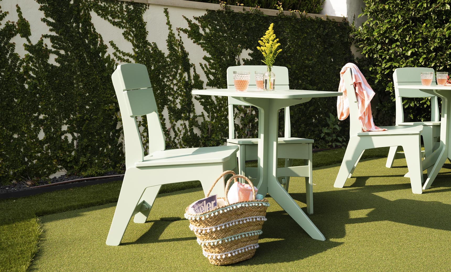 Enjoy a blissful outdoor dining setup with Ledge Lounger’s Mainstay Dining Side Chairs and two light green tables on artificial grass. A woven basket with a magazine and pink water bottle sits below, while a pink cloth drapes over one chair. Yellow flowers add charm to this high-quality scene.