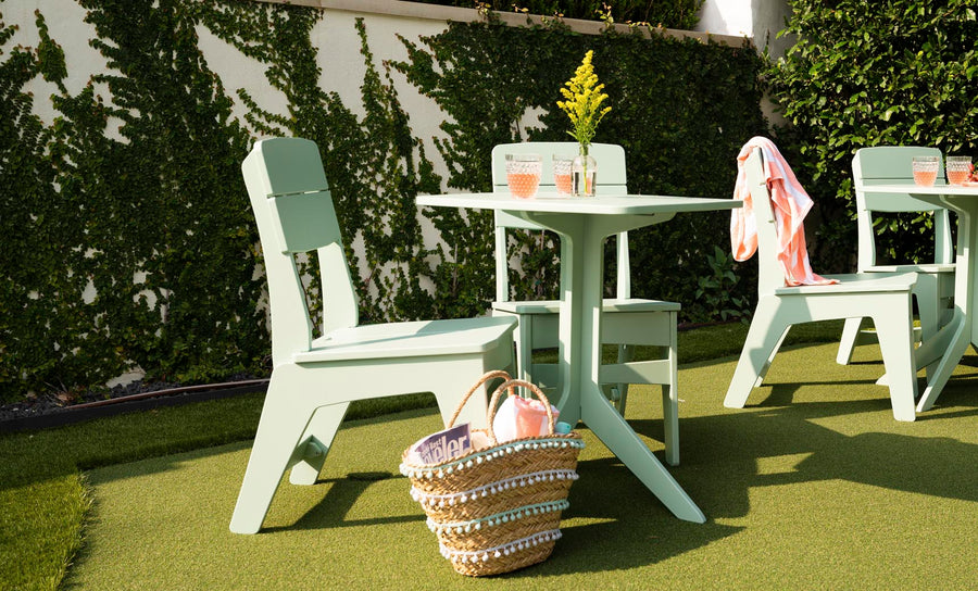 Enjoy a blissful outdoor dining setup with Ledge Lounger’s Mainstay Dining Side Chairs and two light green tables on artificial grass. A woven basket with a magazine and pink water bottle sits below, while a pink cloth drapes over one chair. Yellow flowers add charm to this high-quality scene.
