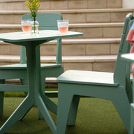 A green patio table and Ledge Lounger Mainstay Dining Side Chairs are placed on artificial grass near stone steps. Two glasses with pink liquid sit on the table atop a striped fabric basket, while a pink and white striped towel is elegantly draped over one chair, ideal for outdoor dining.