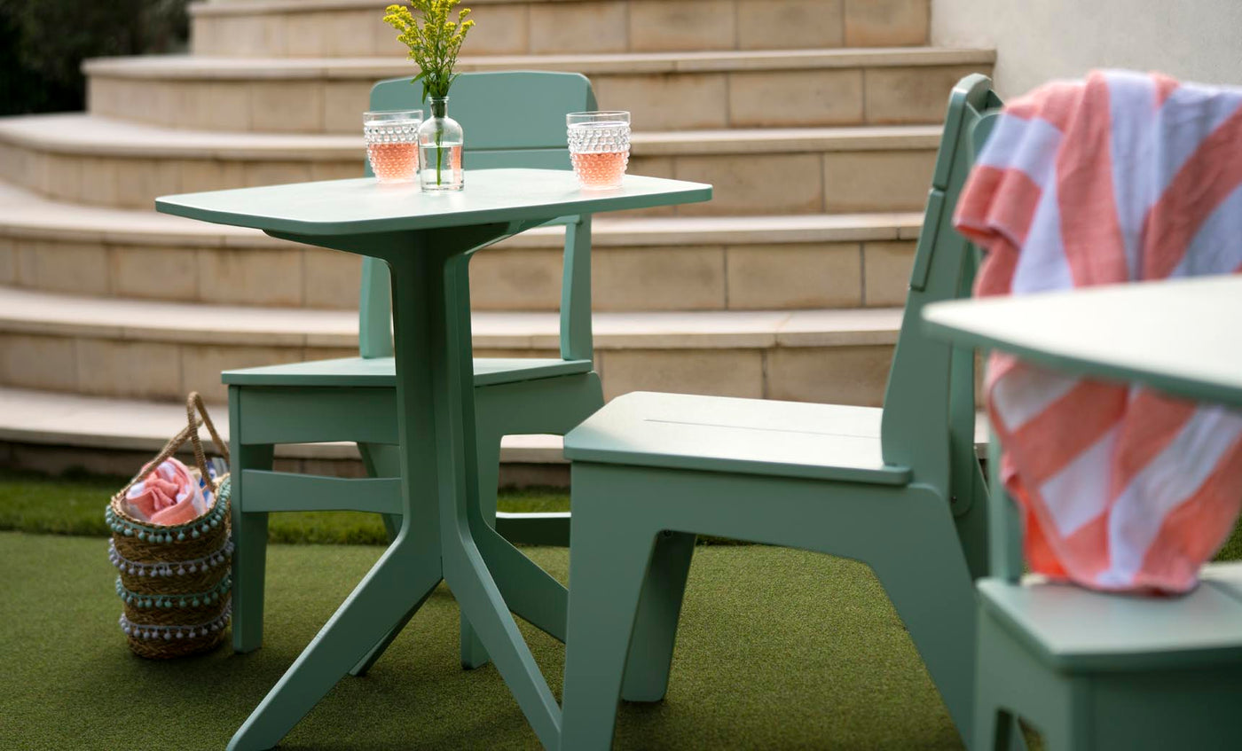 A green patio table and Ledge Lounger Mainstay Dining Side Chairs are placed on artificial grass near stone steps. Two glasses with pink liquid sit on the table atop a striped fabric basket, while a pink and white striped towel is elegantly draped over one chair, ideal for outdoor dining.
