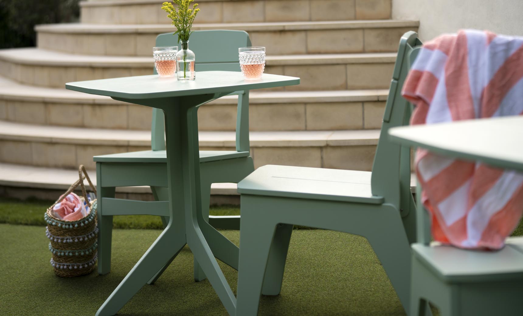 A green patio table and Ledge Lounger Mainstay Dining Side Chairs are placed on artificial grass near stone steps. Two glasses with pink liquid sit on the table atop a striped fabric basket, while a pink and white striped towel is elegantly draped over one chair, ideal for outdoor dining.