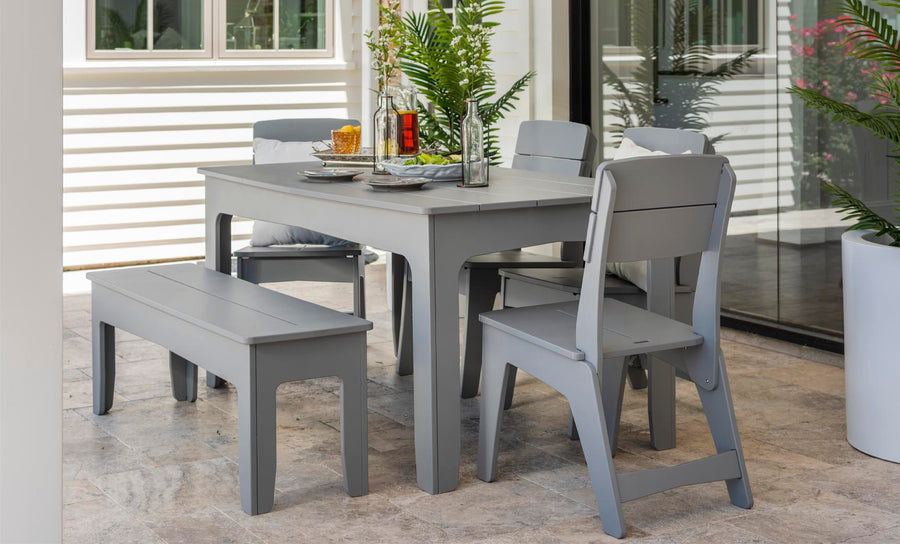 An outdoor dining area showcases the Ledge Lounger Mainstay Rectangular Dining Table in sleek gray, complemented by matching chairs and a bench. The table is adorned with glass bottles, a vase plant, and tableware on a stone-tiled patio adjacent to a glass door with white siding and lush potted plants.