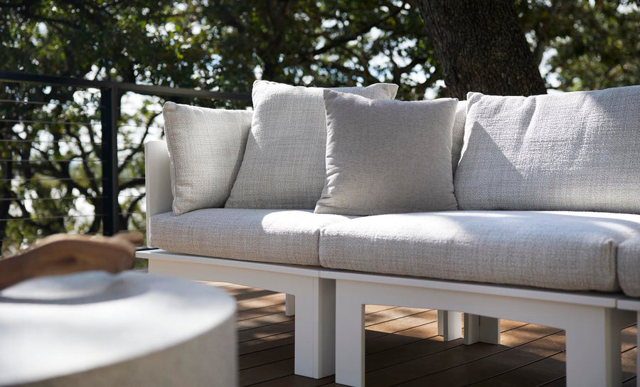 A serene patio features a configurable white sofa with gray cushions on a wooden deck. Nearby, the Ledge Lounger Mainstay Sectional Relaxed Right Armchair blends seamlessly with a table featuring wooden details. Trees and a railing enhance this tranquil garden setting.