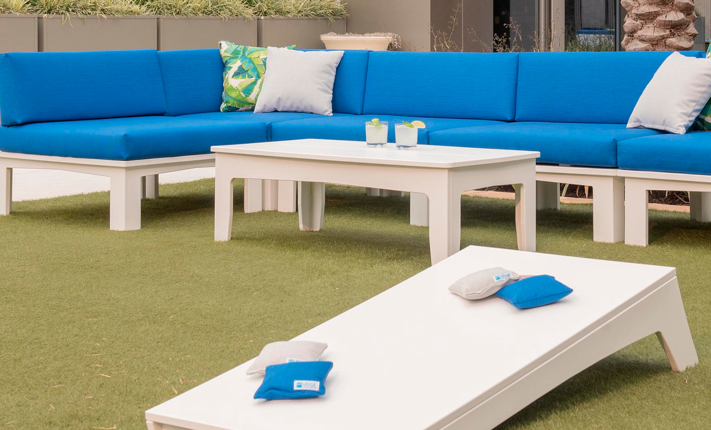 The outdoor seating area showcases blue cushioned couches and a Mainstay rectangular coffee table from Ledge Lounger on lush artificial grass. Two drinks sit invitingly on the table, and a cornhole game with blue and beige bean bags awaits. Green plants form the ideal backdrop for these essentials.
