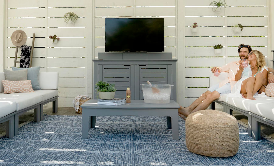 A couple sits on an outdoor sofa enjoying drinks in a cozy patio setup, focused on all-weather durability. The scene includes a TV on a stand, the Mainstay Rectangular Coffee Table with a plant, a large ice bucket, and woven pouf. White slatted walls and geometric rug add modern flair.