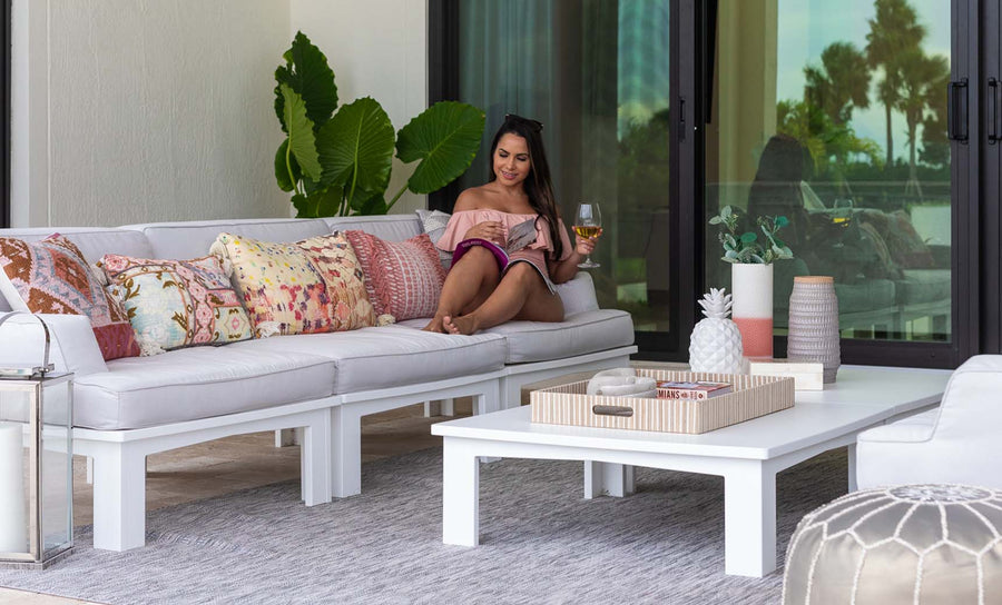 A woman relaxes on a white Ledge Lounger Mainstay Sectional Left Armchair, with a drink and tablet. Colorful pillows from the Mainstay cushion set add vibrancy. Nearby, a white coffee table holds decorative items. Behind her are large green leaves and glass doors reflecting trees.