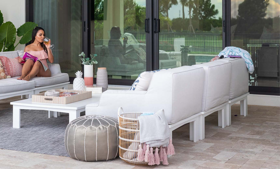 A woman relaxes on the Mainstay Sectional Right Armchair by Ledge Lounger, sipping her drink and perusing a magazine. The white outdoor furniture, with its stylish cushion set and pouf, complements the serene pool view and lush greenery through the glass doors.