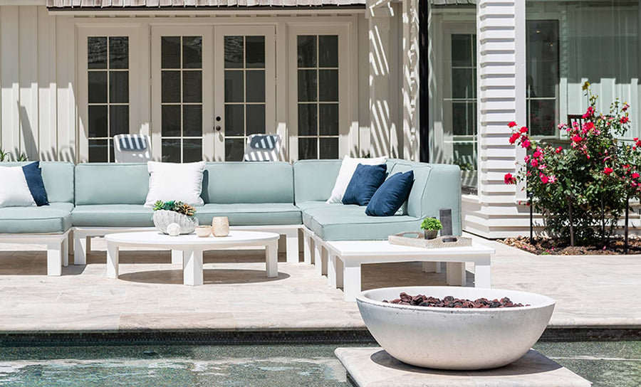 A patio features light blue and white sectional sofas, matching coffee tables, and a Mainstay Sectional Endcap by Ledge Lounger. A round fire pit enhances the area near a pool. Red roses and decorative plants add brightness beside white paneled walls and large glass doors.