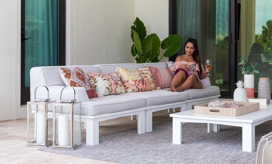 A woman relaxes on the Mainstay Sectional 3 Piece Sofa by Ledge Lounger, adorned with decorative pillows. She sips a drink and holds a tablet on a patio with large glass doors, a rug, and tall candles glowing in lanterns beside her.