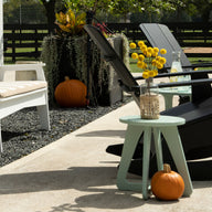 A charming outdoor scene showcases black and white rocking chairs paired with a Mainstay Stool by Ledge Lounger, holding a vase of yellow flowers. Pumpkins sit on the ground as greenery frames the fence-enclosed area under the sunny sky.