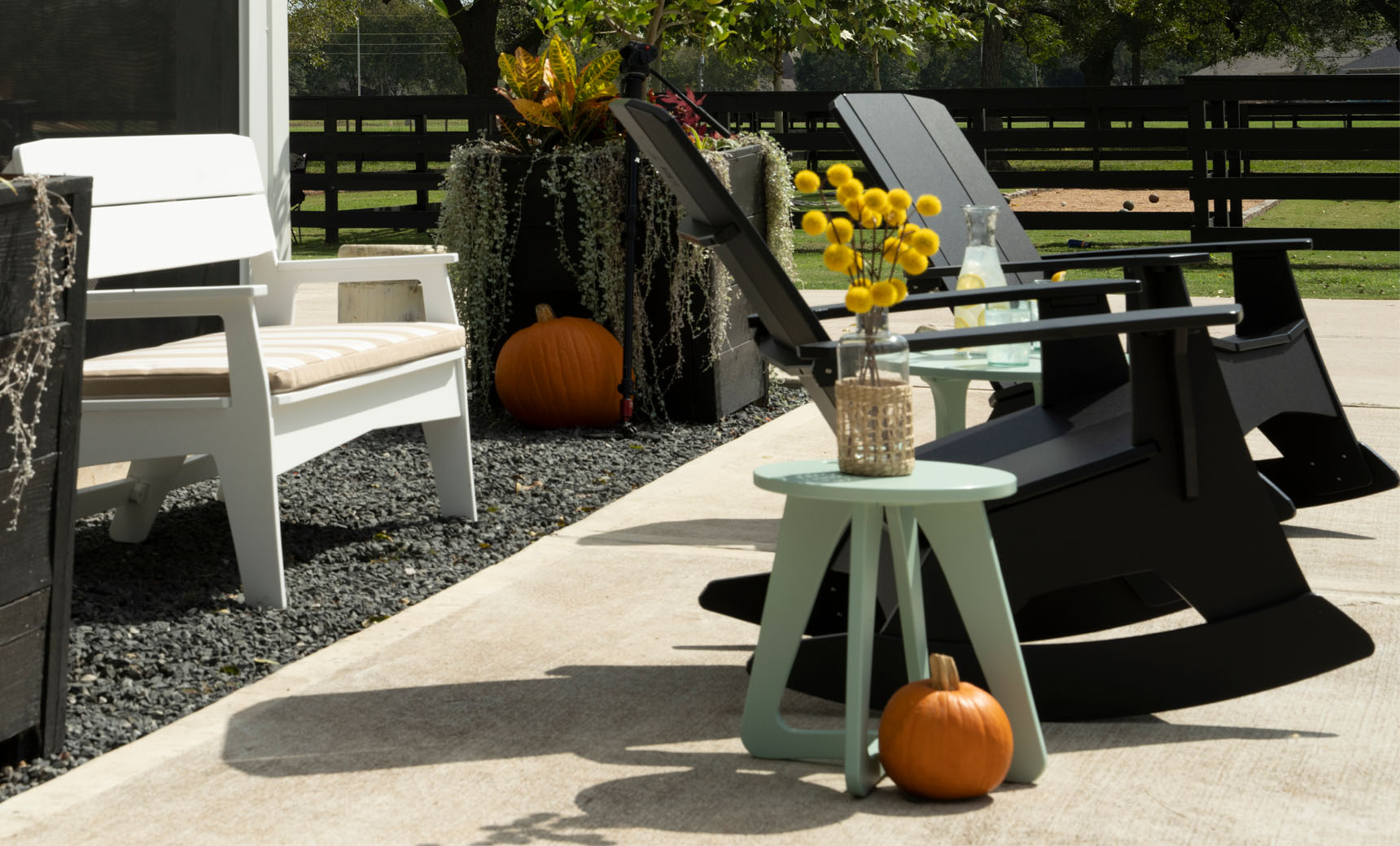 A charming outdoor scene showcases black and white rocking chairs paired with a Mainstay Stool by Ledge Lounger, holding a vase of yellow flowers. Pumpkins sit on the ground as greenery frames the fence-enclosed area under the sunny sky.