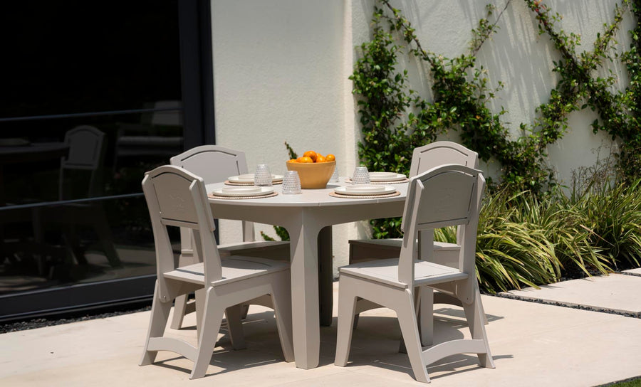 The Mainstay Round Dining Table by Ledge Lounger, complete with an umbrella sleeve, is set on a patio with four beige chairs. It holds plates, glasses, and a bowl of oranges and is placed near a vine-covered wall beside a large window reflecting this serene setting.