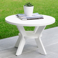 The Mainstay Round Side Table by Ledge Lounger, featuring a cross-legged base, adds elegance to a tiled patio. It supports a small potted succulent and some magazines, while the lush grassy lawn in the background enhances this serene outdoor setting.