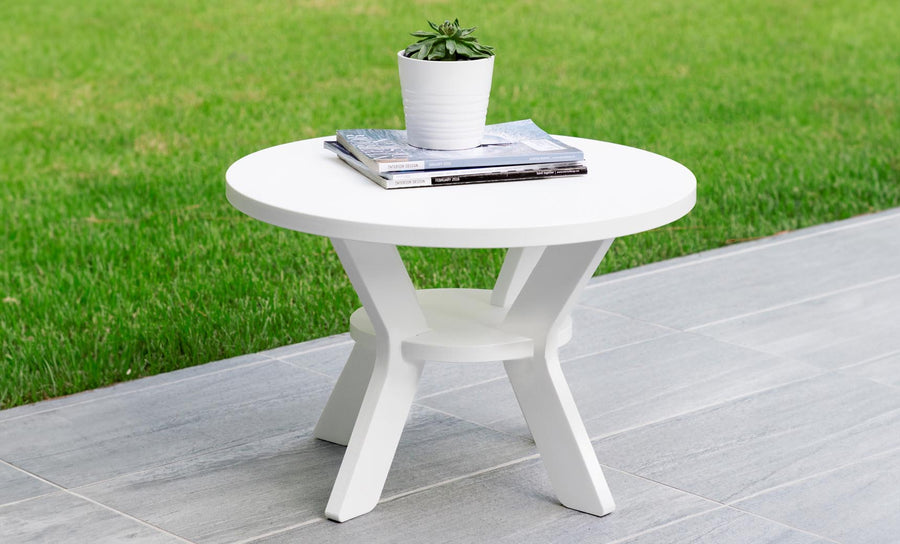 The Mainstay Round Side Table by Ledge Lounger, featuring a cross-legged base, adds elegance to a tiled patio. It supports a small potted succulent and some magazines, while the lush grassy lawn in the background enhances this serene outdoor setting.