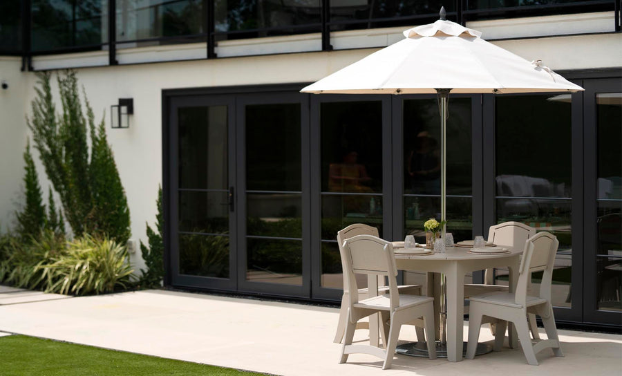 A Ledge Lounger Mainstay Round Dining Table with four matching chairs sits under a large white umbrella. The serene setting, featuring an integrated umbrella sleeve, is on a concrete patio near dark-framed glass doors, with greenery along the wall and grass visible in the foreground.