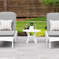 Two gray cushioned lounge chairs with striped pillows sit on a tiled patio, showcasing a minimalistic design. A Ledge Lounger Mainstay Round Side Table between them holds two glasses of white wine. The outdoor space is framed by lush bushes, trees, and a wooden fence.