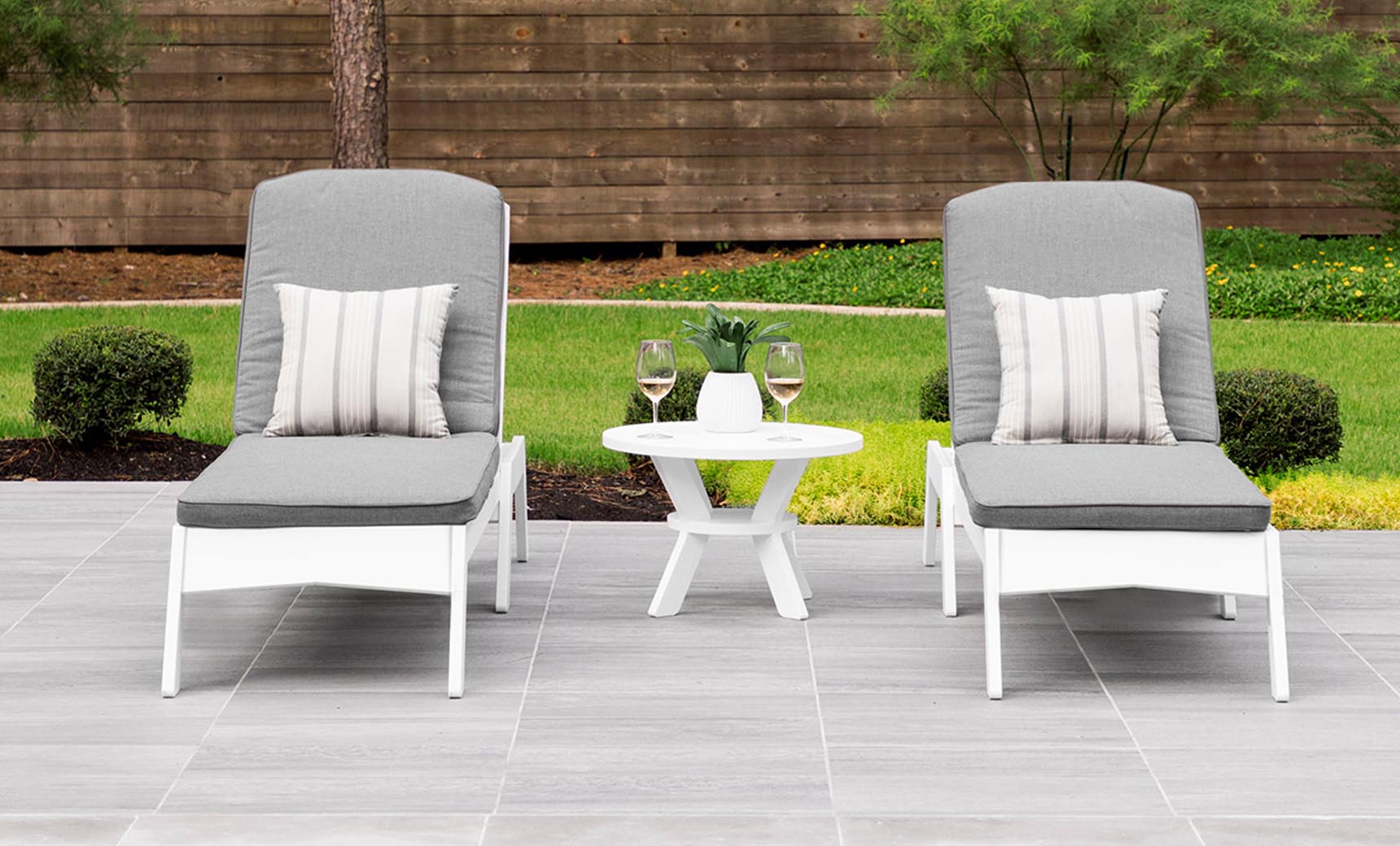 Two gray cushioned lounge chairs with striped pillows sit on a tiled patio, showcasing a minimalistic design. A Ledge Lounger Mainstay Round Side Table between them holds two glasses of white wine. The outdoor space is framed by lush bushes, trees, and a wooden fence.