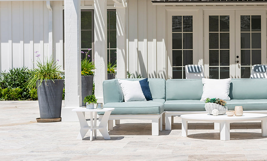 A patio features a light blue sectional sofa with white cushions, paired with a Mainstay Square Side Table in white holding decorative items. A tall planter with greenery sits on the left, and the background displays a white house with double glass doors.