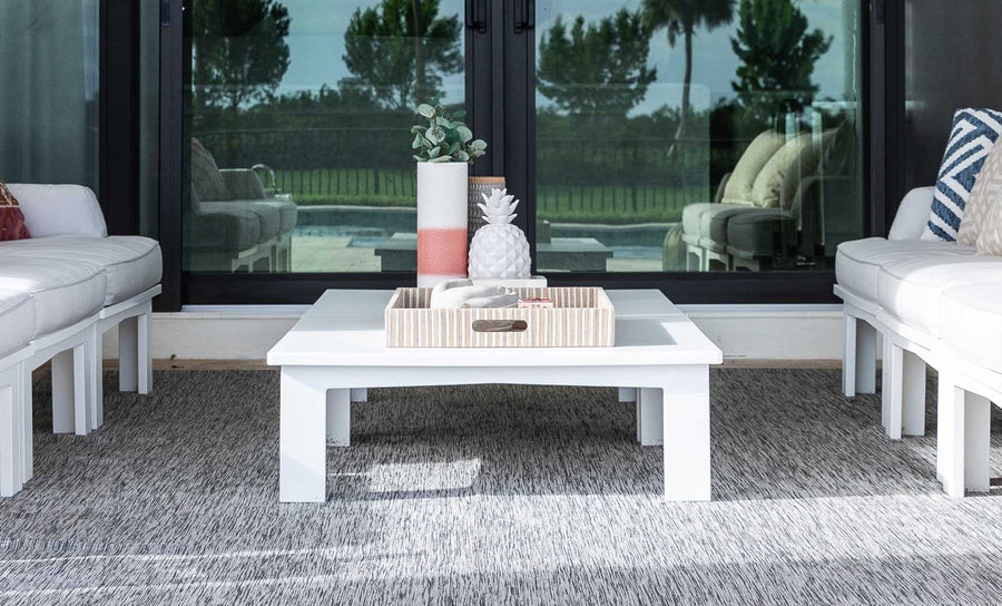 A modern outdoor seating area with white cushioned sofas, a Mainstay Square Coffee Table by Ledge Lounger on a gray rug, featuring a decorative tray with a vase and pineapple ornament. Glass sliding doors in the background reveal trees and a pool outside.
