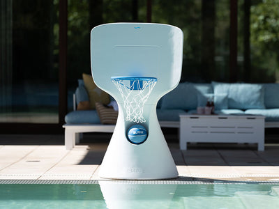 A modern, blue and white basketball hoop is positioned poolside. The background shows a patio area with a cushioned outdoor sofa and a coffee table, partially shaded and reflecting sunlight.