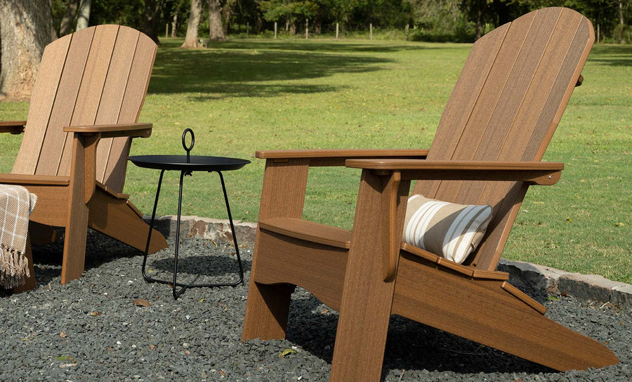 Two wooden Adirondack chairs with a Ledge Lounger 10x18 rectangular throw pillow rest on a pebble-covered area in a grassy yard. A small black metal table stands between them, with a plaid blanket draped over one arm of the chair. Trees and an expansive lawn are visible in the background.