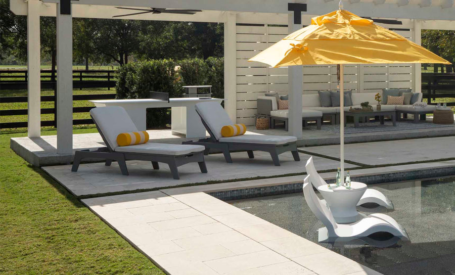 An outdoor patio features two lounge chairs with Ledge Loungers Round Bolster Throw Pillows beside a pool. A floating pool chair with a yellow umbrella is in the water, while a sectional sofa and tall wooden fence are surrounded by greenery in the background.
