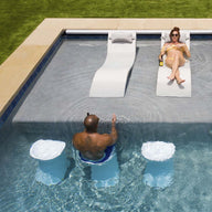 A man and a woman relax in a pool; shes on a submerged chair, wearing sunglasses and sipping her drink. He sits facing her on an underwater Ledge Lounger Signature Barstool Bar Height. The lush grass surrounding the pool creates an ideal spot for outdoor entertaining.
