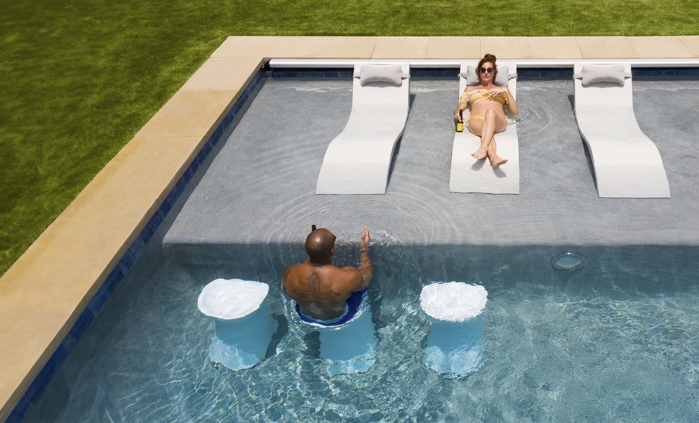 A man and a woman relax in a pool; shes on a submerged chair, wearing sunglasses and sipping her drink. He sits facing her on an underwater Ledge Lounger Signature Barstool Bar Height. The lush grass surrounding the pool creates an ideal spot for outdoor entertaining.