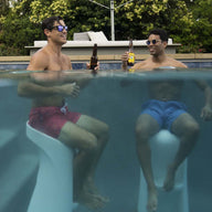 Two men, wearing sunglasses and swim trunks, relax poolside on Signature Barstools by Ledge Lounger, half-submerged in the water while holding beer bottles. The lush backdrop of trees enhances the perfect outdoor entertaining vibe.
