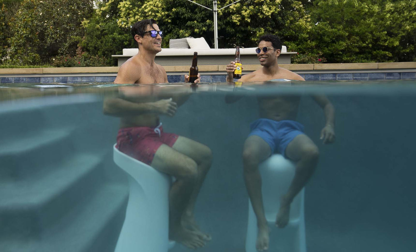 Two men, wearing sunglasses and swim trunks, relax poolside on Signature Barstools by Ledge Lounger, half-submerged in the water while holding beer bottles. The lush backdrop of trees enhances the perfect outdoor entertaining vibe.