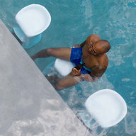 A person in blue swim shorts sits on a Ledge Lounger Signature Barstool Bar Height at the poolside bar, holding a drink. Clear water reveals two white stools nearby. Perfect for outdoor entertaining, they appear relaxed and partially submerged in this unique aquatic setting.