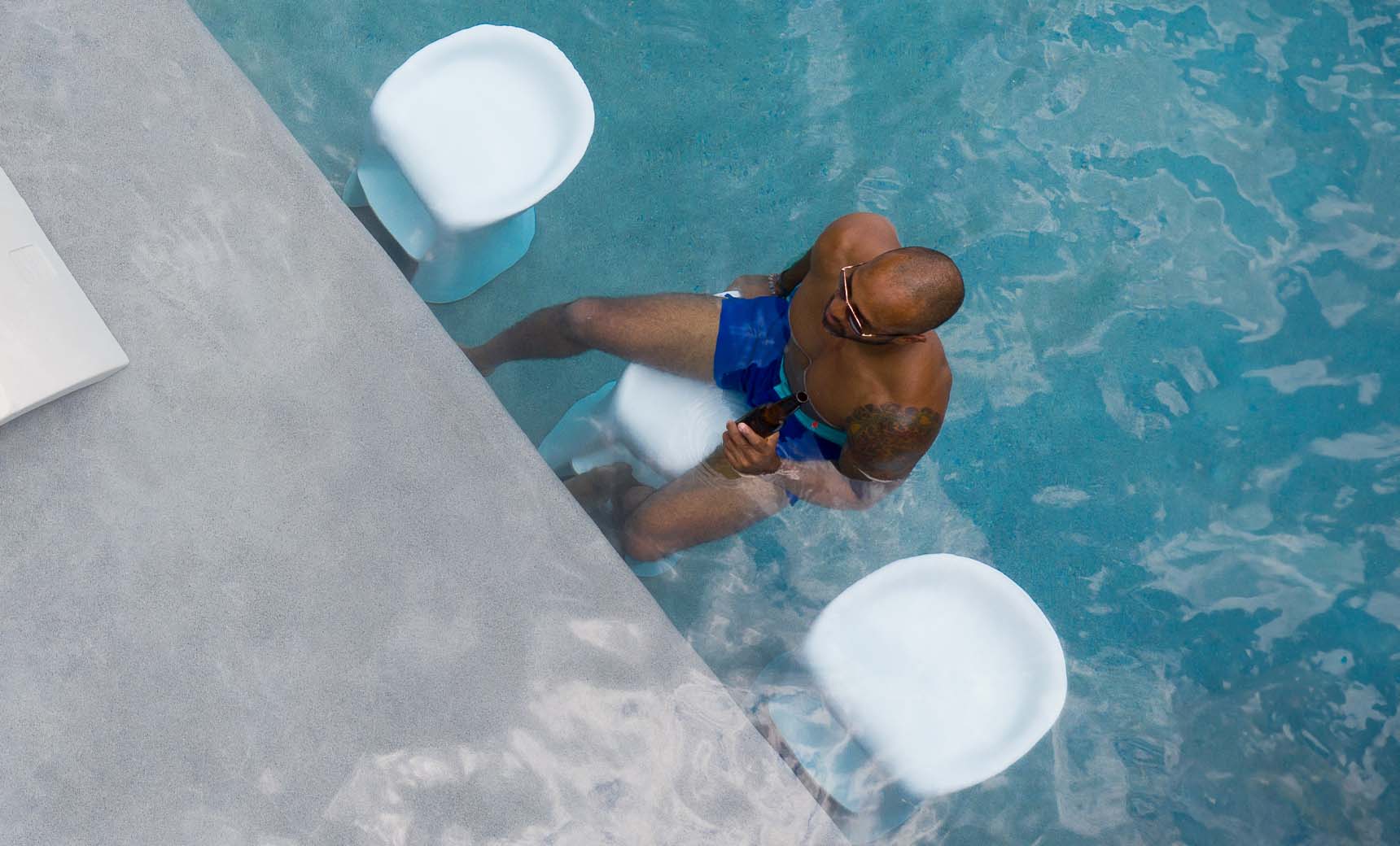 A person in blue swim shorts sits on a Ledge Lounger Signature Barstool Bar Height at the poolside bar, holding a drink. Clear water reveals two white stools nearby. Perfect for outdoor entertaining, they appear relaxed and partially submerged in this unique aquatic setting.