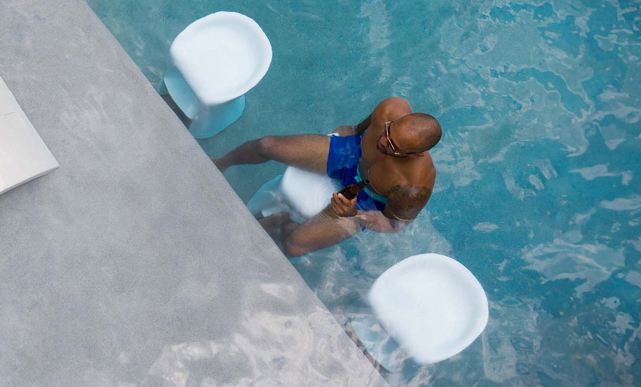 A person in blue swim shorts sits on a Ledge Lounger Signature Barstool Bar Height at the poolside bar, holding a drink. Clear water reveals two white stools nearby. Perfect for outdoor entertaining, they appear relaxed and partially submerged in this unique aquatic setting.