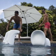 Two shirtless men wearing sunglasses and blue-red swim trunks playfully push white Signature Barstool Bar Height chairs by Ledge Lounger into the pool. An open white umbrella and lush green trees create an ideal outdoor entertaining backdrop.