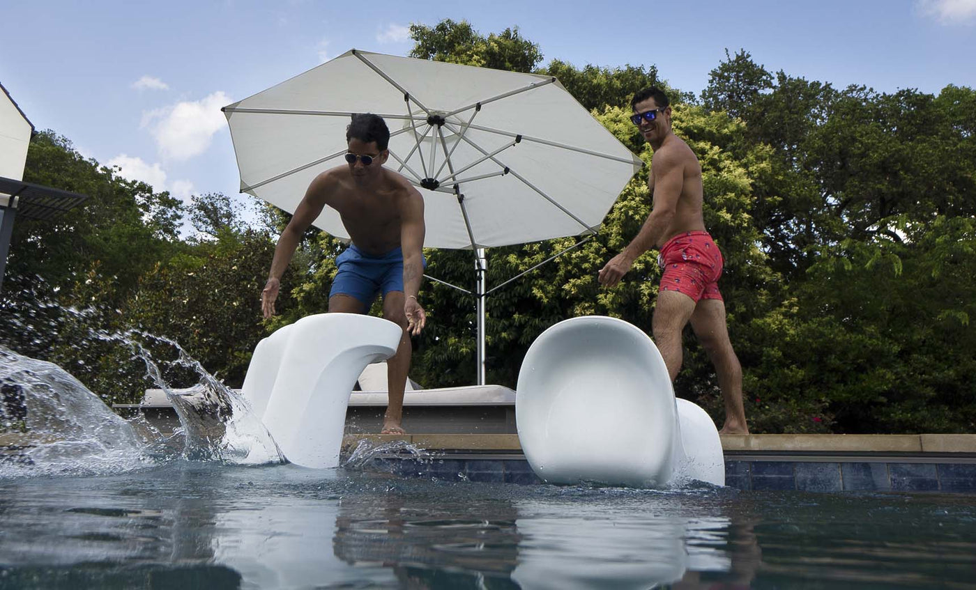 Two shirtless men wearing sunglasses and blue-red swim trunks playfully push white Signature Barstool Bar Height chairs by Ledge Lounger into the pool. An open white umbrella and lush green trees create an ideal outdoor entertaining backdrop.
