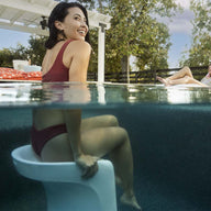 A woman in a red swimsuit sits on a submerged Ledge Lounger Signature Barstool Bar Height in the pool, smiling at the sky and capturing outdoor enjoyment. Nearby, someone relaxes by the pool under a pergola framed by trees.