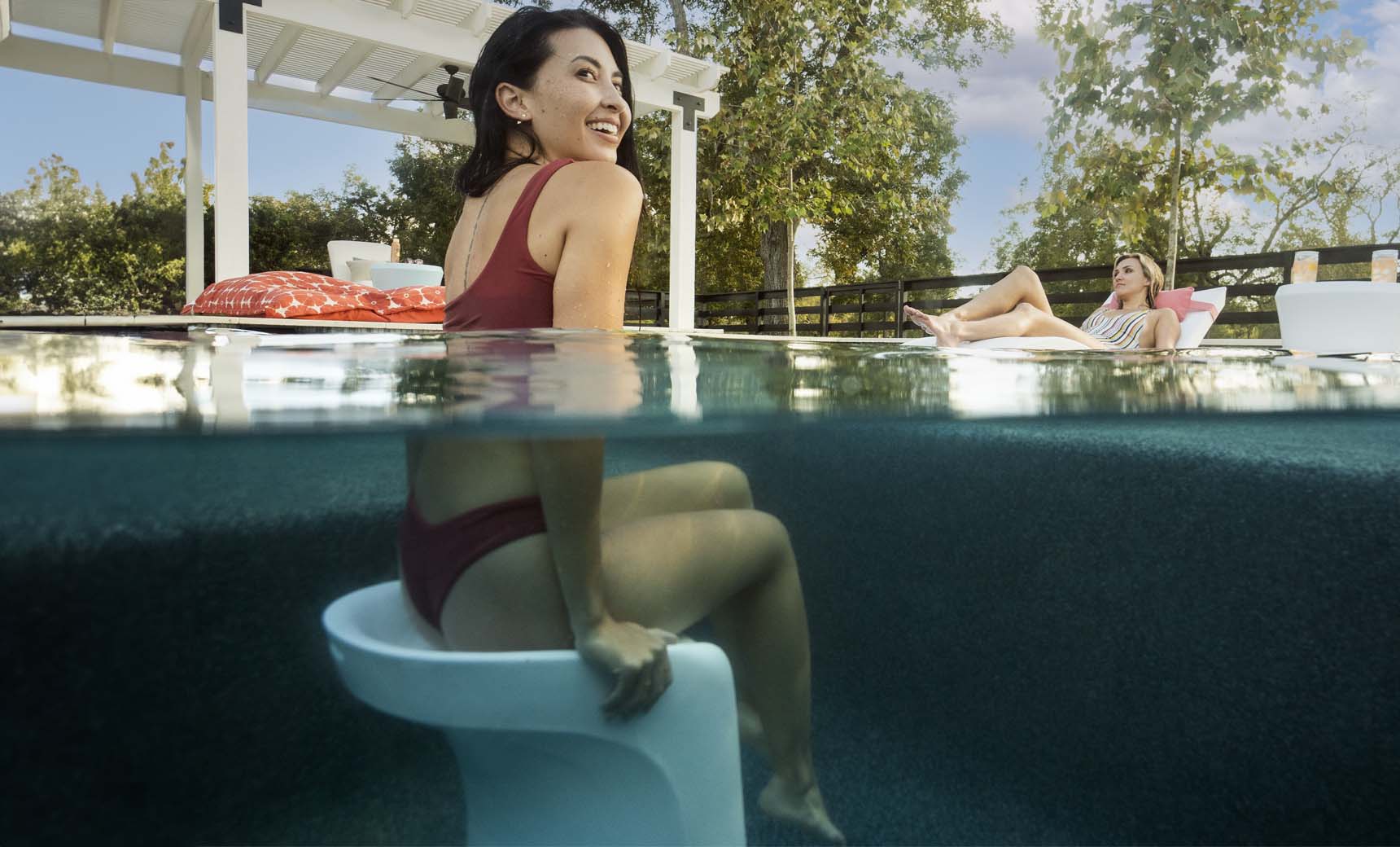 A woman in a red swimsuit sits on a submerged Ledge Lounger Signature Barstool Bar Height in the pool, smiling at the sky and capturing outdoor enjoyment. Nearby, someone relaxes by the pool under a pergola framed by trees.