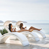 Three people in swimwear enjoy the sun on modern, white Signature Chaise loungers by Ledge Lounger overlooking a calm sea. Two recline with drinks while another kneels at the pools edge, drink in hand, all captured through a vivid media viewing window in this serene setting.