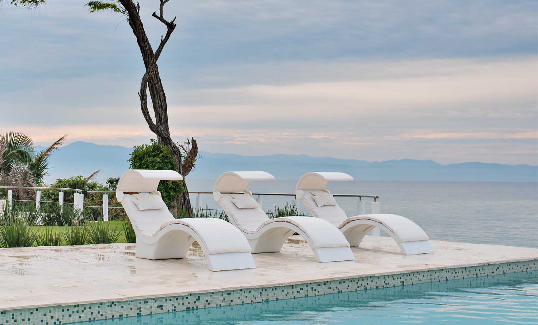 Three Ledge Lounger Signature Chaise Shades are arranged on a poolside patio, overlooking serene ocean waters. A solitary tree and lush greenery enhance the background, with clouds touching distant mountains at the horizon.