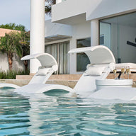 By a modern pool, three Ledge Lounger Chaise chairs are gracefully submerged, each equipped with a Signature Chaise Shade and plush pillows. Nearby, a small table holds two glasses and a bottle. A contemporary white building and lush greenery provide the perfect backdrop.
