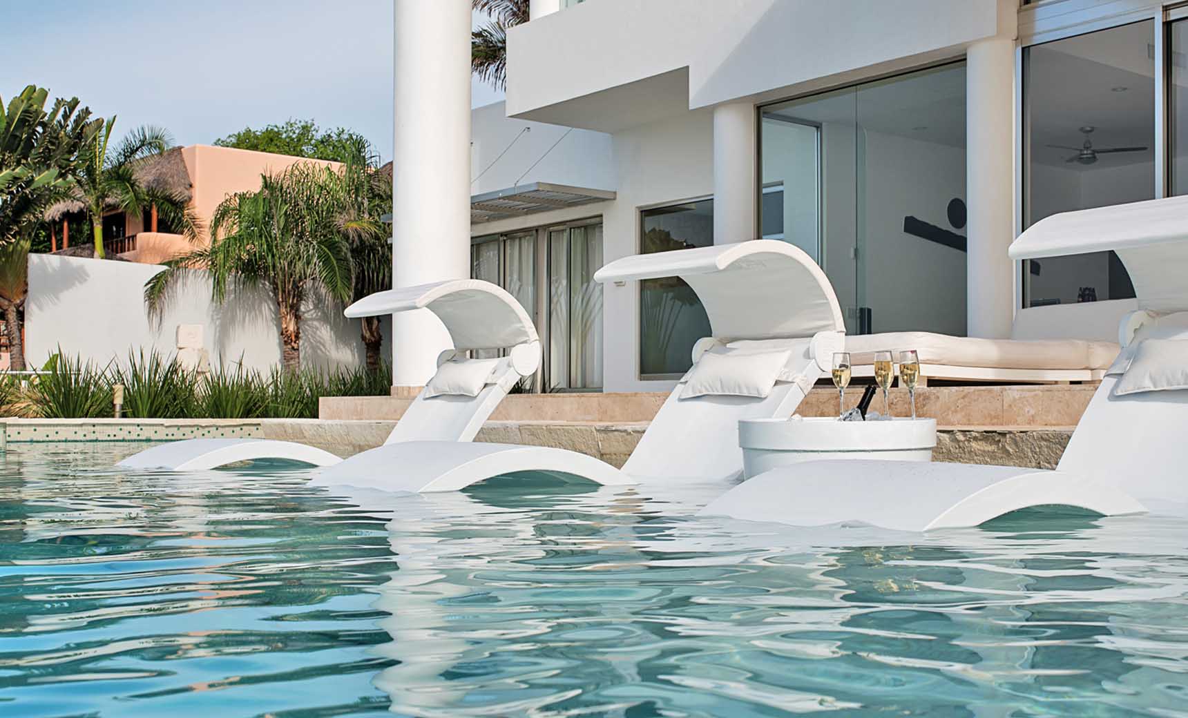 By a modern pool, three Ledge Lounger Chaise chairs are gracefully submerged, each equipped with a Signature Chaise Shade and plush pillows. Nearby, a small table holds two glasses and a bottle. A contemporary white building and lush greenery provide the perfect backdrop.