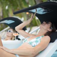 Two people relax on Ledge Loungers Signature Chaises in a pool. The foreground figure, under the Signature Chaise Shade, sports sunglasses and a printed bikini while using a touchscreen device. Nearby, another person enjoys a drink surrounded by palm trees.