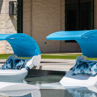Two Ledge Lounger Signature Chaise Shade lounge chairs with blue canopies and patterned pillows sit in shallow pool water, perfect for sun relaxation. A stone building and green grass are visible in the background.