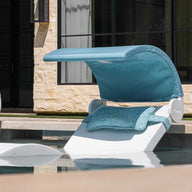 Poolside view of two Ledge Lounger Signature Chaises with teal cushions and adjustable Signature Chaise Shades partially submerged in water near a stone building with large media windows, showcasing modern relaxation and style.