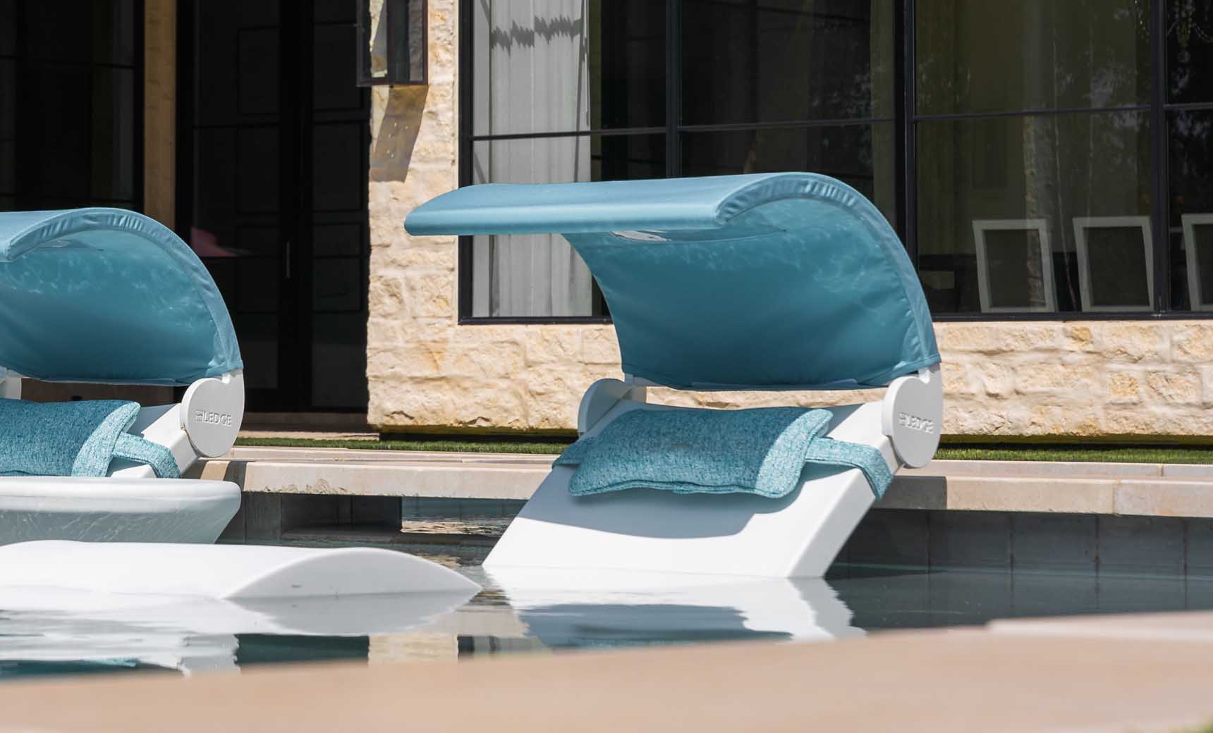 Poolside view of two Ledge Lounger Signature Chaises with teal cushions and adjustable Signature Chaise Shades partially submerged in water near a stone building with large media windows, showcasing modern relaxation and style.