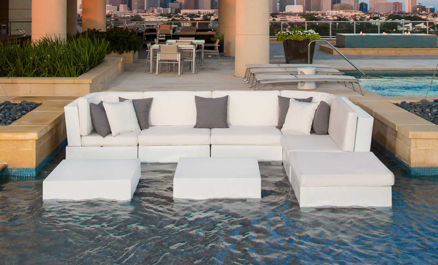A modern outdoor lounge features a white sectional set and Ledge Loungers Signature Coffee Table in a shallow pool. A dining table with chairs against the skyline backdrop offers an exquisite view, highlighting our Signature Collection.