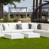 The Signature Collection outdoor setup includes a white sectional sofa with gray pillows on grass, modern white cube tables, and tall potted plants. A wicker pod structure against the city skyline completes this poolside scene. The Signature Coffee Table is by Ledge Lounger.