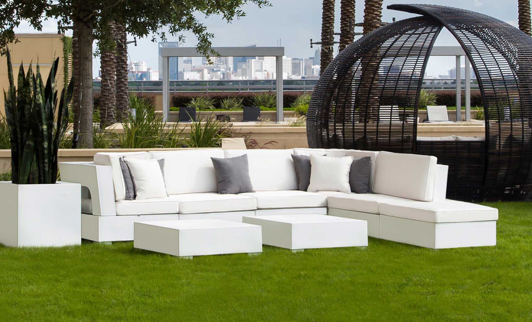 The Signature Collection outdoor setup includes a white sectional sofa with gray pillows on grass, modern white cube tables, and tall potted plants. A wicker pod structure against the city skyline completes this poolside scene. The Signature Coffee Table is by Ledge Lounger.
