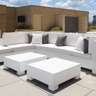A modern patio boasts white modular sofas and a Signature Coffee Table by Ledge Lounger from the Signature Collection. Grey and white cushions provide contrast, complemented by large potted plants. The setup overlooks a pool under a blue sky, ideal for poolside relaxation essentials.