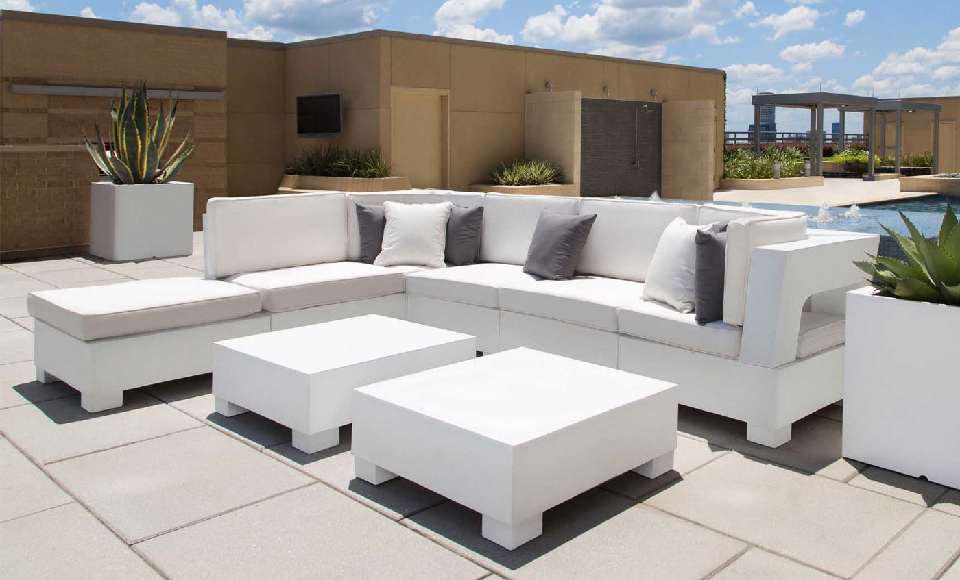 A modern patio boasts white modular sofas and a Signature Coffee Table by Ledge Lounger from the Signature Collection. Grey and white cushions provide contrast, complemented by large potted plants. The setup overlooks a pool under a blue sky, ideal for poolside relaxation essentials.